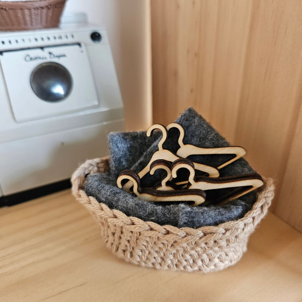 A small crocheted basket holding a stack of miniature wooden dollhouse hangers on a soft gray felt lining, placed on a wooden shelf with a miniature washing machine in the background.