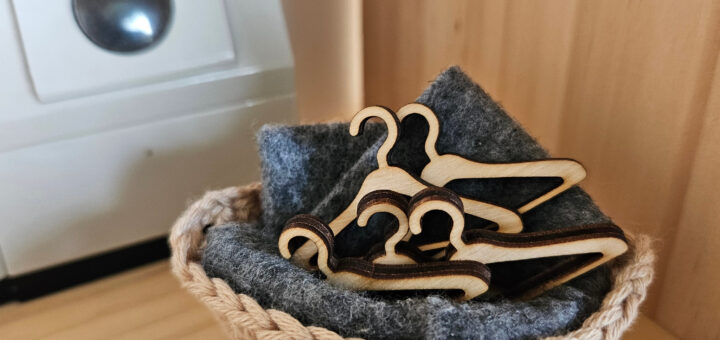 A small crocheted basket holding a stack of miniature wooden dollhouse hangers on a soft gray felt lining, placed on a wooden shelf with a miniature washing machine in the background.