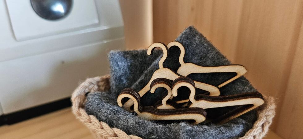 A small crocheted basket holding a stack of miniature wooden dollhouse hangers on a soft gray felt lining, placed on a wooden shelf with a miniature washing machine in the background.